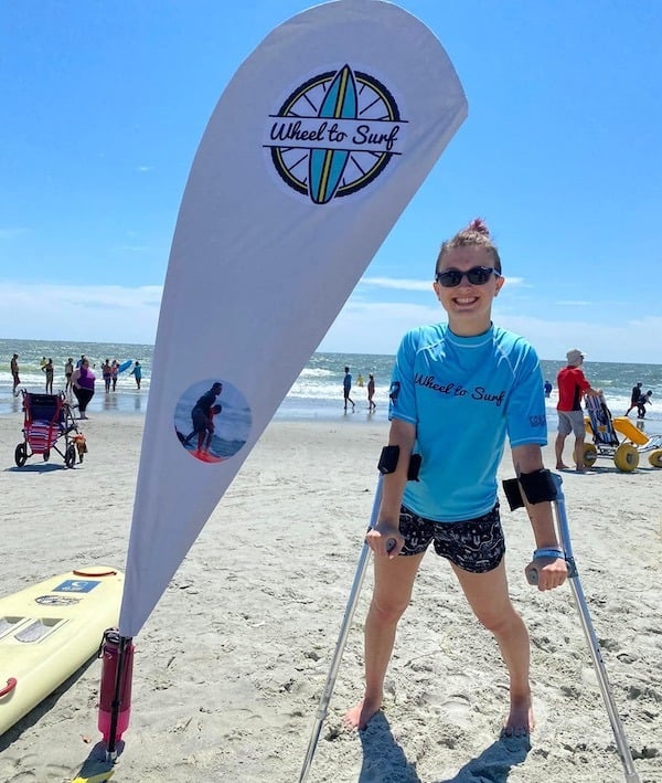 girl with sunglasses and crutches standing next to Wheel to Surf banner on beach